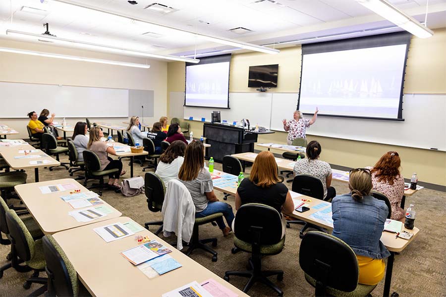 More than a dozen teachers sit in a large classroom as an instructor discusses a topic.