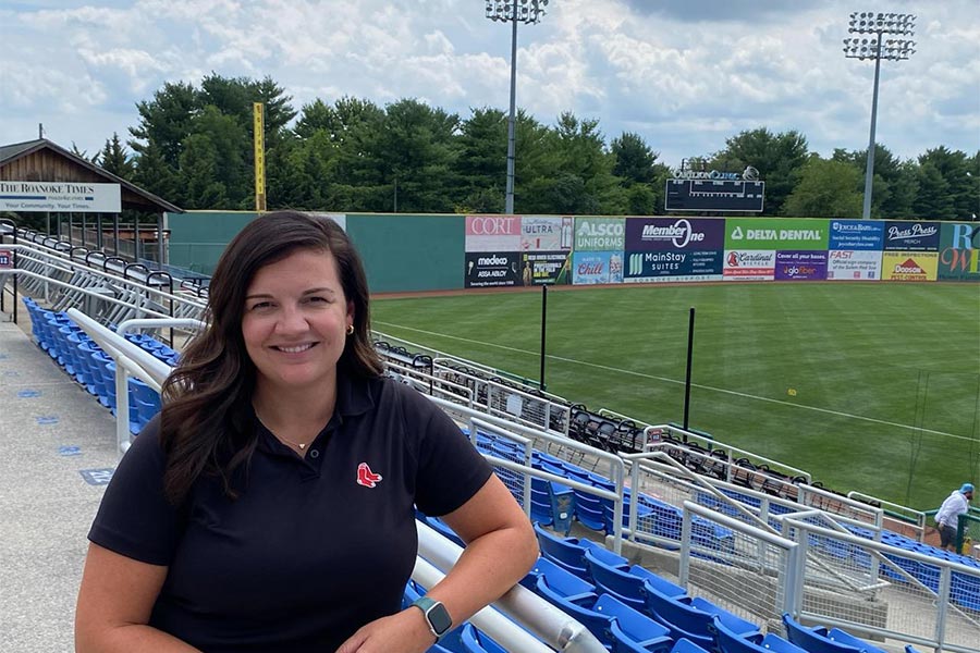 A woman stands smiling, with a ballpark and blue sky behind her.