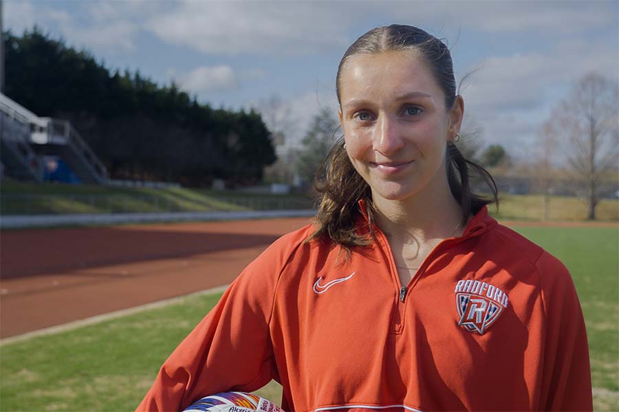 A marketing graduate and Radford University soccer star stands on the playing field with a ball under her arm.