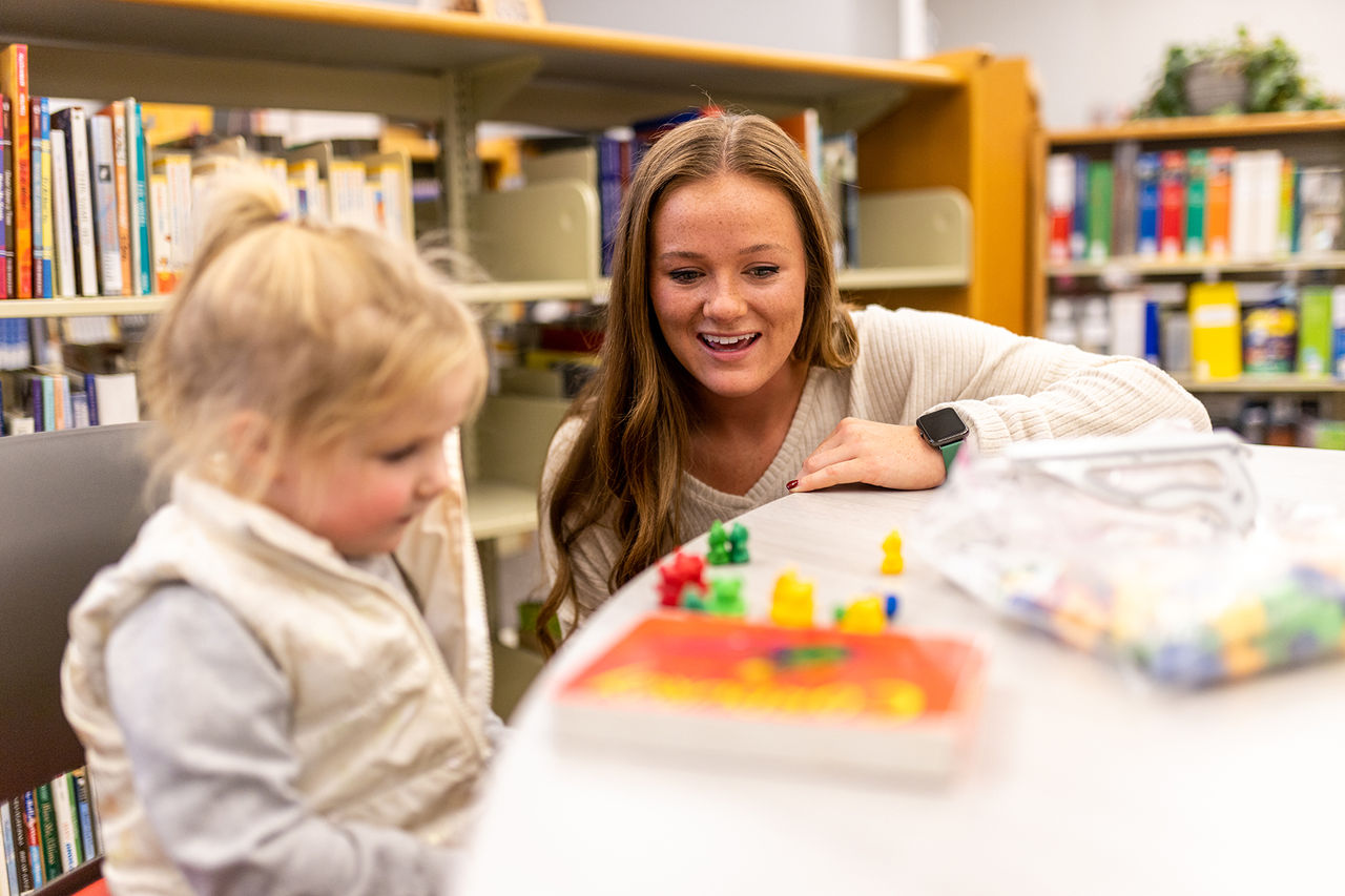 A woman leaning on a table next to a small child who has a few small colorful items in front of her. 