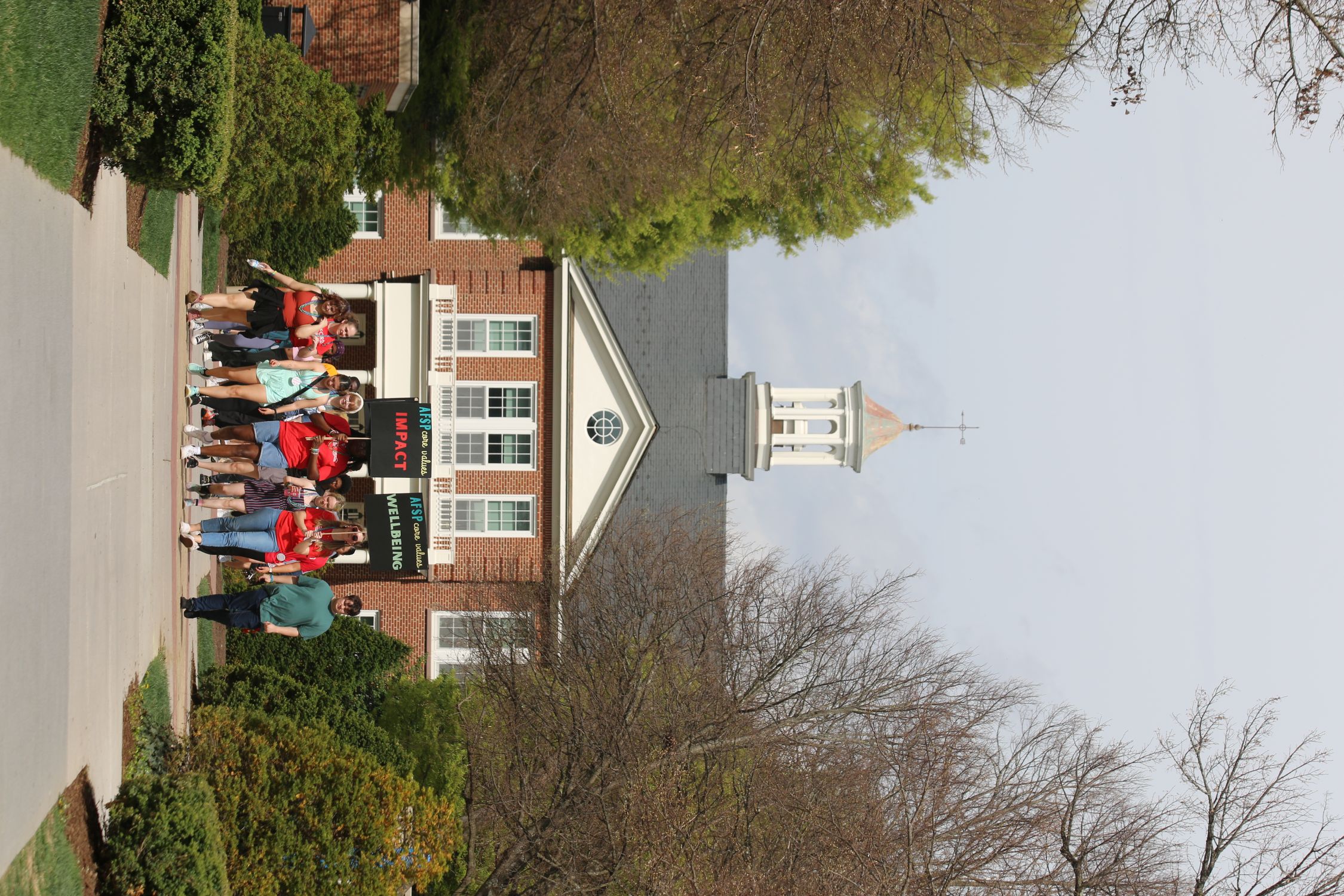 A group of people walks along a tree‑lined pathway toward a red‑brick campus building with white trim and a cupola on the roof. The building has banners hanging above the entrance with the words “IMPACT” and “WELLBEING.” Trees frame the walkway on both sides, and the scene appears to be on a clear day.