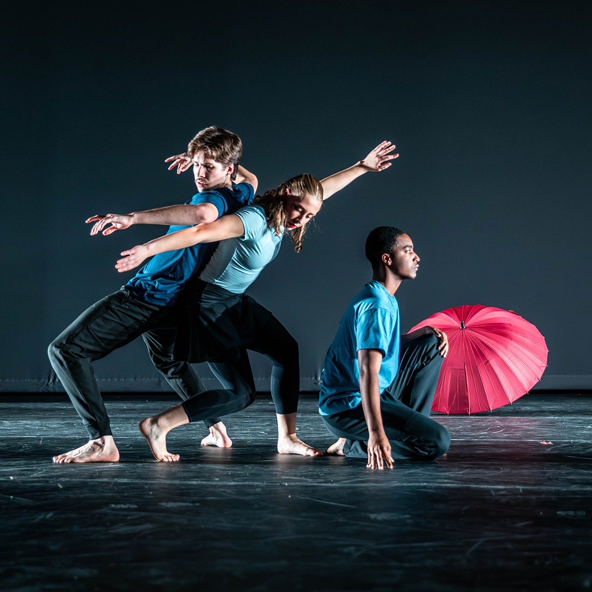Three dancers in black pants and blue shirts perform on a dark stage with a red umbrella.