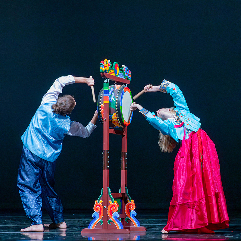 Dancers in traditional Korean clothing strike a drum while bending over backwards.