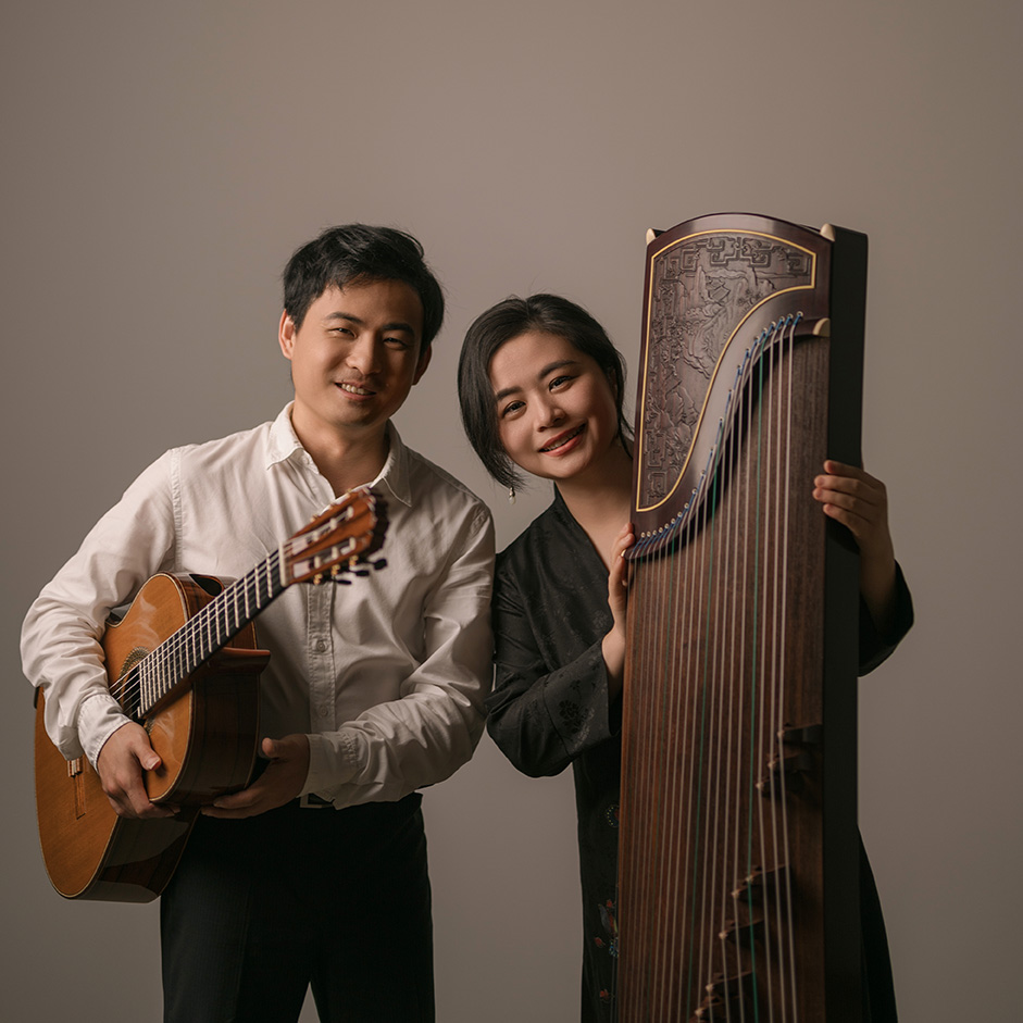 A man, Bin Hu, and woman, Jing Xai, stand together smiling. Bin Hu holds a guitar. Jing Xai holds a guzheng.