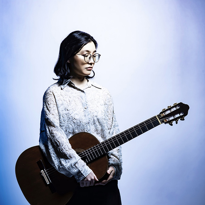 A woman (Hao Yang) stands in front of a bluish backdrop with a guitar strapped over her shoulder.