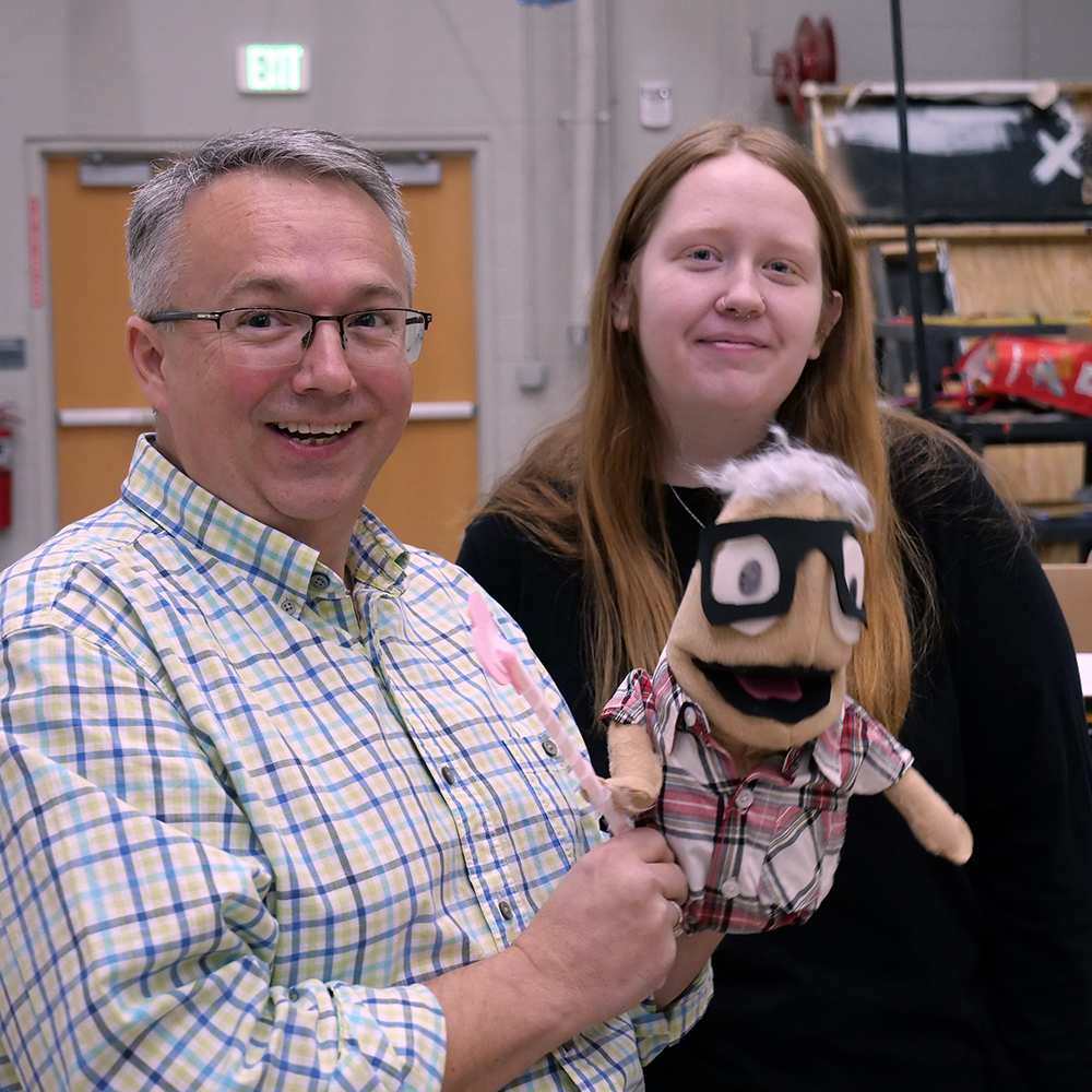 A man smiling in the foreground holds a puppet with a young lady stand smiling behind him.