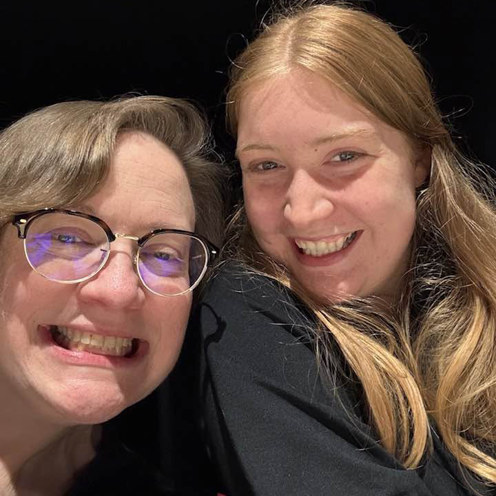 Two smiling women, Kate Burnham-Hull and Sarah Lindsey Merriman, lean into the center of the frame smiling against a black background.