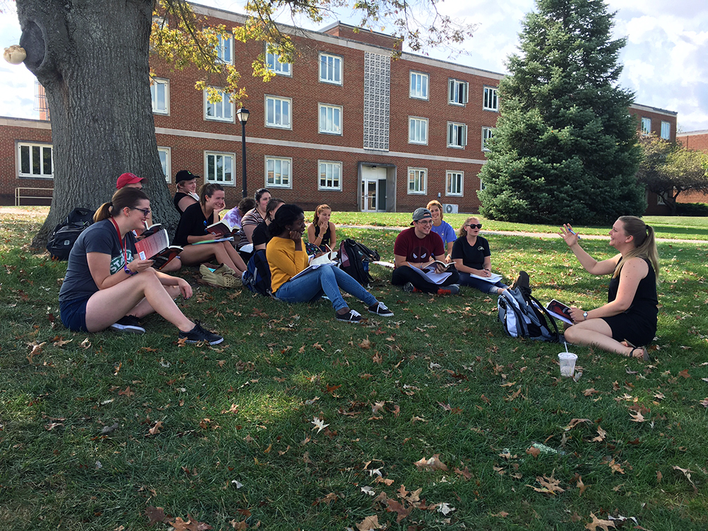 A group of college students sit beneath a large tree on Radford University's campus as their professor, Molly Hood, conducts class.